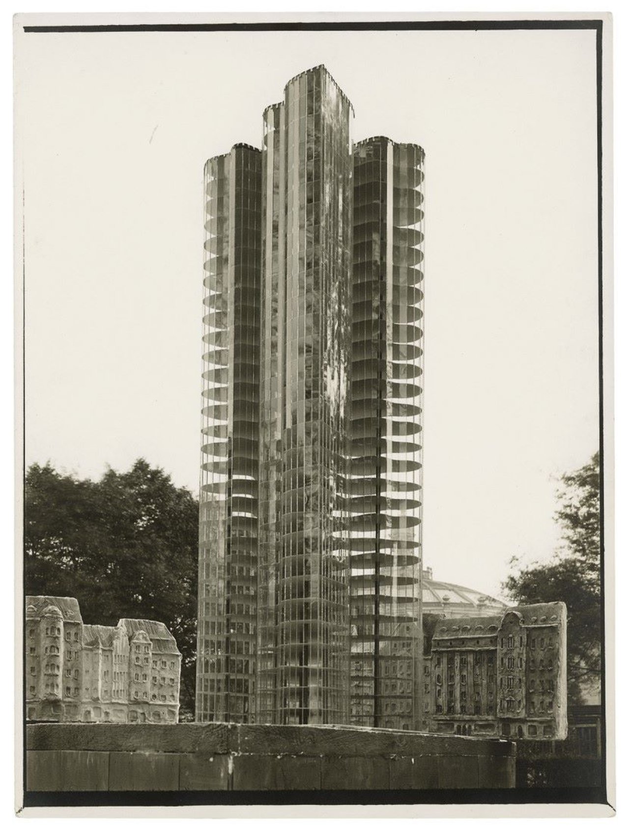 Auf diesem Bild ist das folgende Kunstwerk zu sehen: Curt Rehbein. Mies van der Rohe, Gläserner Wolkenkratzer, Modell mit Staffage von Oskar Herzog vor dem Gebäude des ehemaligen Kolonialmuseums. Nicht ausgeführtes Projekt von 1922.