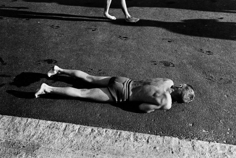 Auf diesem Bild ist das folgende Kunstwerk zu sehen: Will McBride. „Junge wärmt sich am Asphalt neben dem Schwimmbecken, Berlin (Süd)“. 1956.
