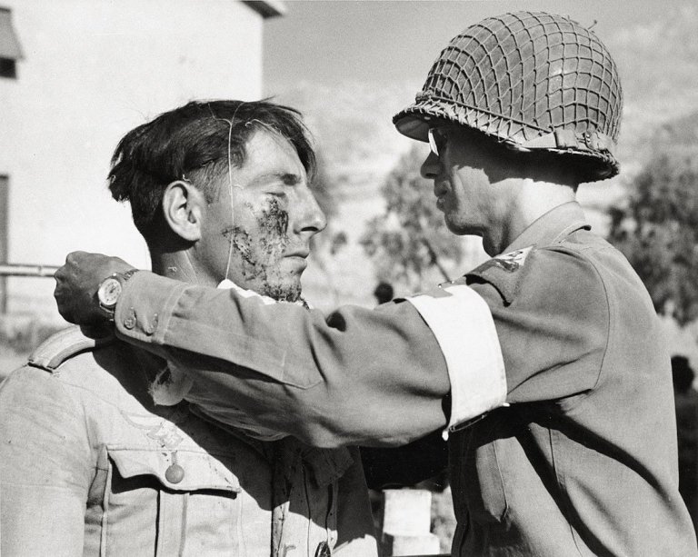 Auf diesem Bild ist das folgende Kunstwerk zu sehen: Robert Capa (d.i. Endre Ernő Friedmann). Erste Hilfe eines amerikanischen Militärarztes für einen verletzten deutschen Soldaten, Sizilien. 1943.