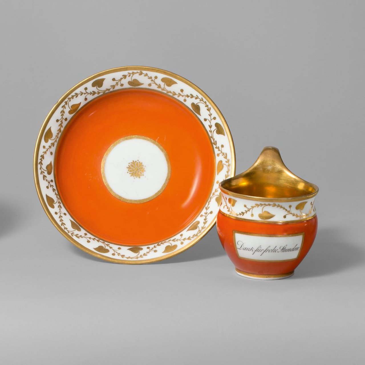 Auf diesem Bild ist das folgende Kunstwerk zu sehen:  KPM Königliche Porzellan-Manufaktur Berlin. Gedenktasse Etrurische Form mit Sinnspruch „Dank für frohe Stunden“ auf orange-rotem Fond und reicher Vergoldung. Um 1800.