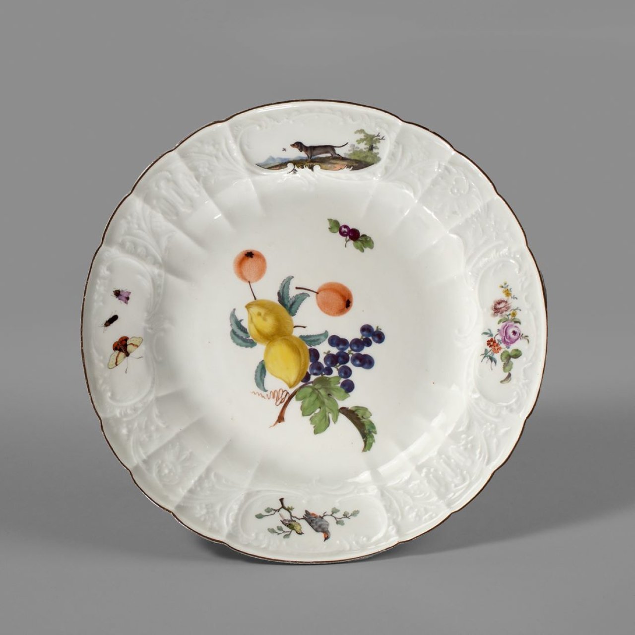Auf diesem Bild ist das folgende Kunstwerk zu sehen:  Porzellanmanufaktur Meissen. Teller mit Dulong-Relief, Früchten und brauner Randkante. Um 1765.